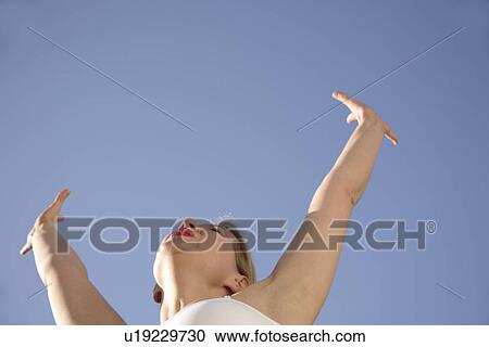 Stock Image - Woman stretching out arms in front of blue sky, low angle view. Fotosearch