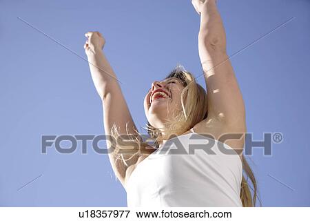 Young woman cheering with arms raised in front of blue sky, view from below View Large Photo Image Stock Photo - Young woman cheering with arms raised in front of blue sky, view from below. Fotosearch