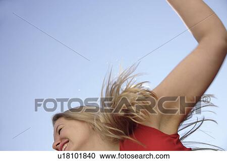 Stock Image - Young woman dancing and cheering with arms stretched out in front of blue sky, low angle view. Fotosearch