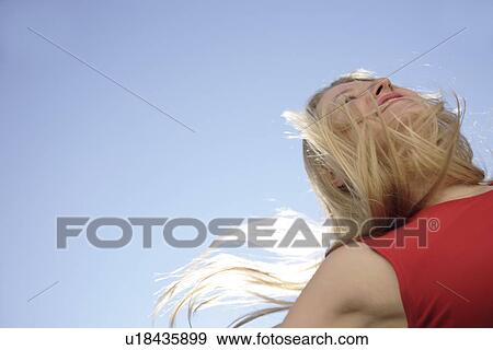 Stock Photo - Young woman throwing her long blond hair in front of blue sky, low angle view. Fotosearch