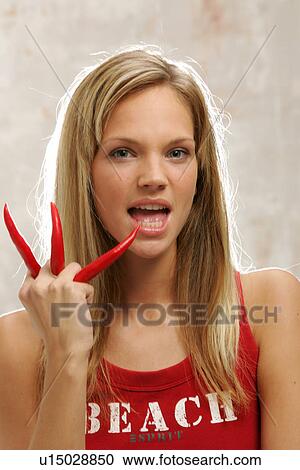 Stock Image - Young woman with long blond hair holding three chili peppers, portrait. Fotosearch