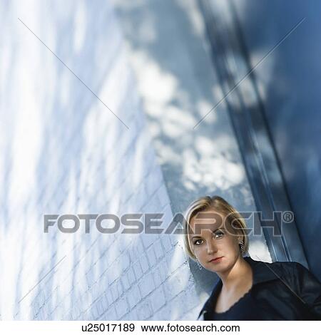 Stock Photo - Blonde woman standing against sun dappled building looking at viewer. Fotosearch