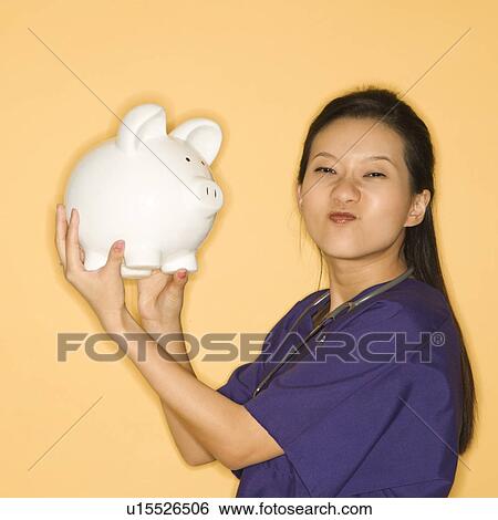 Female doctor holding piggy bank against yellow background scrunching nose and looking at viewer. View Large Photo Image Stock Photograph - Female doctor holding piggy bank against yellow background scrunching nose and looking at viewer.. Fotosearch