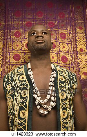 Stock Image - Low angle portrait of mid-adult man wearing embroidered African vest and beads. Fotosearch