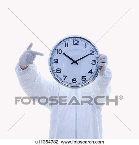 Stock Image - Man in biohazard suit holding clock in front of face pointing to clock with finger standing against white background.. Fotosearch