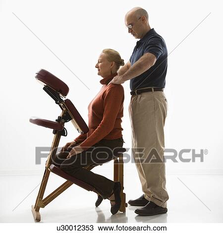 Stock Image - Massage therapist massaging shoulders of woman sitting in massage chair. Fotosearch