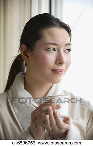 Stock Image - Mid adult woman in bathrobe drinking coffee and looking out window. Fotosearch