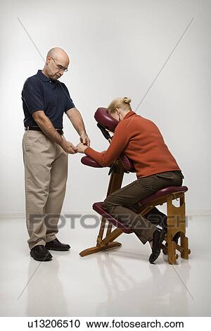 Stock Image - Middle-aged male massage therapist massaging hands of middle-aged woman sitting in massage chair. Fotosearch