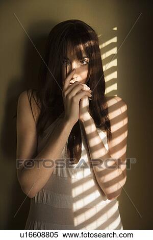 Stock Photography - Portrait of young woman standing against wall in sunlight with hands over mouth. Fotosearch