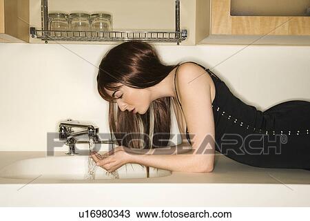 Stock Image - Pretty young woman lying on kitchen counter drinking water from faucet.. Fotosearch