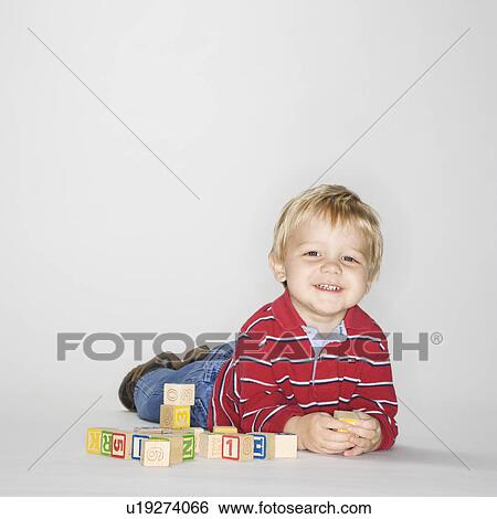 Stock Photograph - Studio portrait of young boy laying on stomach propped up on elbows playing with toy blocks. Fotosearch
