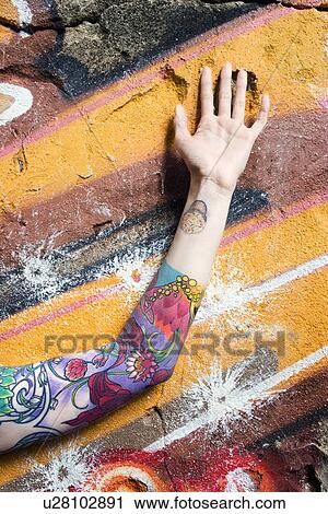 Stock Image - Tattooed woman's arm against graffiti covered wall. Fotosearch