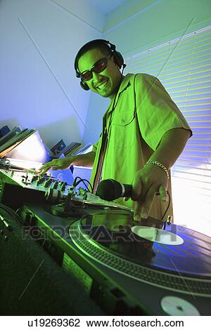 Young adult DJ wearing sunglasses behind mixing equipment with hand on record turntable looking at viewer smiling View Large Photo Image Stock Image - Young adult DJ wearing sunglasses behind mixing equipment with hand on record turntable looking at viewer smiling. Fotosearch