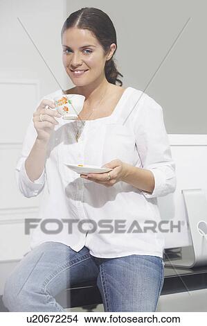 Picture - Smiling woman sitting on desk drinking tea.. Fotosearch