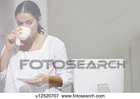 Stock Photo - Woman drinking tea at computer desk.. Fotosearch