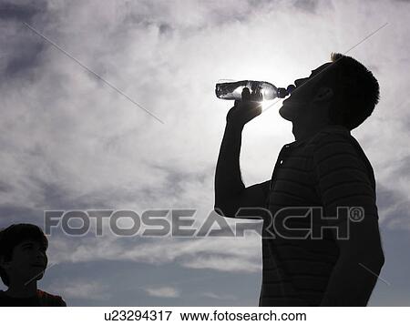 Stock Photo - Man drinking water from bottle, son (8-10) looking on (backlit, lens flare). Fotosearch