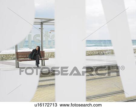Stock Image - Woman waiting at tram stop viewed through some large lettering on glass shelter. Alicante, Spain.. Fotosearch