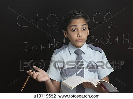 Stock Image - Close-up of a boy holding a pencil and a book in front of a blackboard. Fotosearch