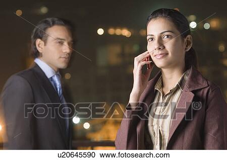 Stock Image - Close-up of a businesswoman talking on a mobile phone with a young man standing in the background. Fotosearch