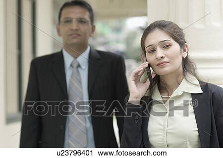 Stock Image - Close-up of a businesswoman talking on a mobile phone with a businessman standing behind her. Fotosearch