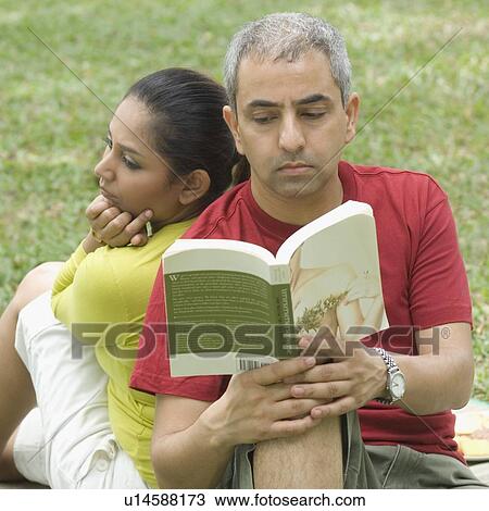 Stock Image - Close-up of a mid adult man reading a book with a mid adult woman sitting behind him. Fotosearch