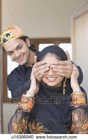 Stock Image - Close-up of a young man covering his wife's eyes from behind. Fotosearch
