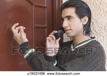 Stock Photograph - Close-up of a young man talking on a mobile phone and knocking on a door. Fotosearch