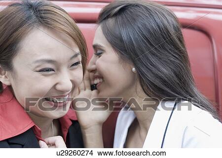 Picture - Close-up of a young woman whispering into another woman's ear. Fotosearch