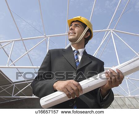 Stock Photo - Low angle view of a male architect holding a blueprint at a construction site. Fotosearch