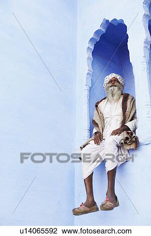 Stock Image - Low angle view of a senior man sitting on an arch window, Pushkar, Rajasthan, India. Fotosearch