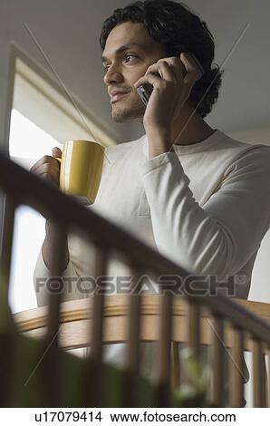 Low angle view of a young man talking on a mobile phone and holding a coffee cup View Large Photo Image Picture - Low angle view of a young man talking on a mobile phone and holding a coffee cup. Fotosearch