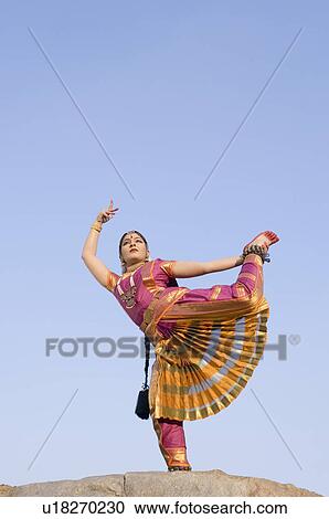 Stock Image - Low angle view of a young woman performing Bharatnatyam on a rock. Fotosearch