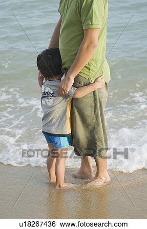 Stock Photograph - Mid adult man standing with his son on the beach. Fotosearch