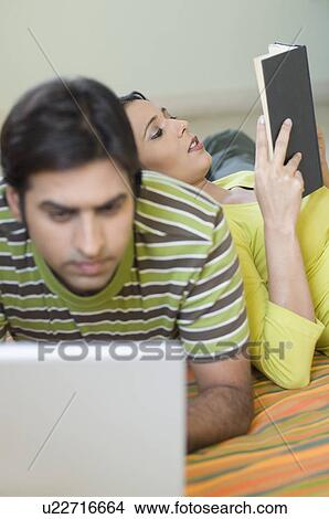 Picture - Mid adult man using a laptop with a young woman leaning on his back and reading a book. Fotosearch