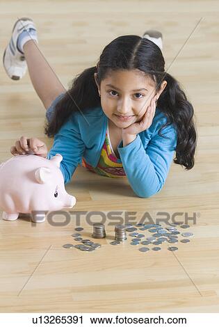 Stock Image - Portrait of a girl lying on the floor and putting coins into a piggy bank. Fotosearch