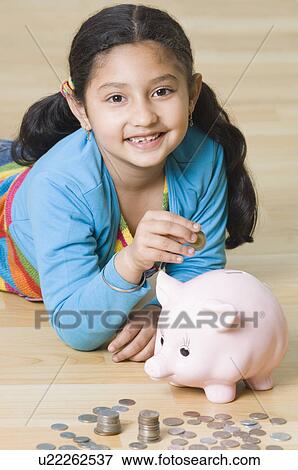 Stock Photo - Portrait of a girl lying on the floor and putting coins into a piggy bank. Fotosearch