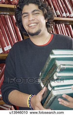 Stock Image - Portrait of a young man carrying a stack of books in a library and smiling. Fotosearch