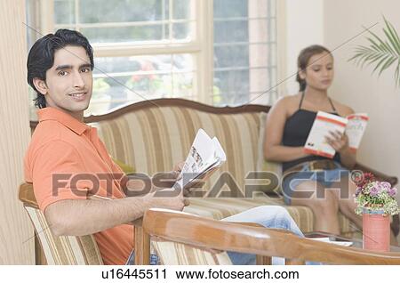 Stock Image - Portrait of a young man holding a newspaper with a young woman reading a book in the background. Fotosearch