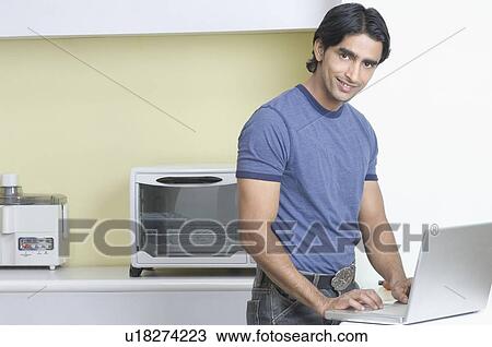 Stock Image - Portrait of a young man standing in the kitchen and using a laptop. Fotosearch