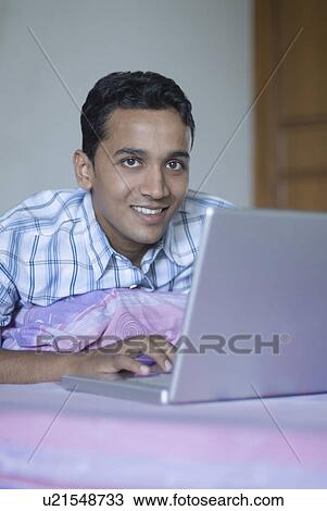 Stock Image - Portrait of a young man using a laptop on a bed. Fotosearch