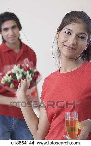 Portrait of a young woman holding a champagne flute with a young man holding a bouquet of flowers behind her View Large Photo Image Picture - Portrait of a young woman holding a champagne flute with a young man holding a bouquet of flowers behind her. Fotosearch