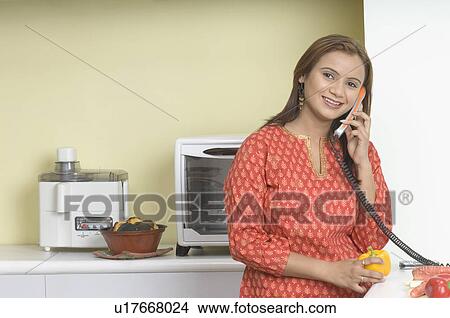 Picture - Portrait of a young woman using a telephone in the kitchen. Fotosearch