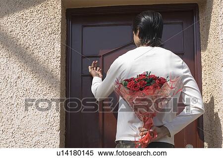Stock Photography - Rear view of a man hiding a bouquet of flowers behind his back and knocking the door. Fotosearch