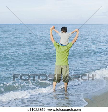 Stock Photo - Rear view of a mid adult man carrying his son on his shoulders on the beach. Fotosearch