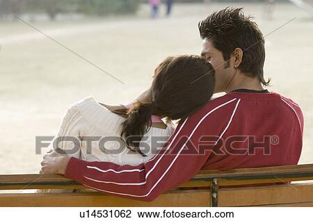 Stock Image - Rear view of a young couple sitting together on a park bench. Fotosearch