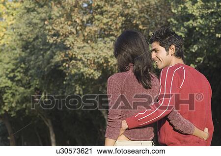 Stock Image - Rear view of a young couple walking with their arms around each other. Fotosearch