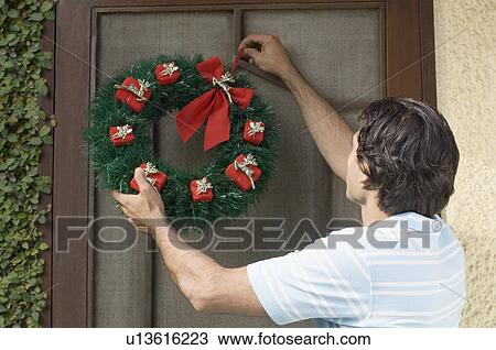 Stock Image - Rear view of a young man hanging wreath on a door. Fotosearch