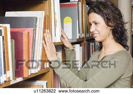 Stock Image - Side profile of a female college student selecting a book from a library shelf. Fotosearch