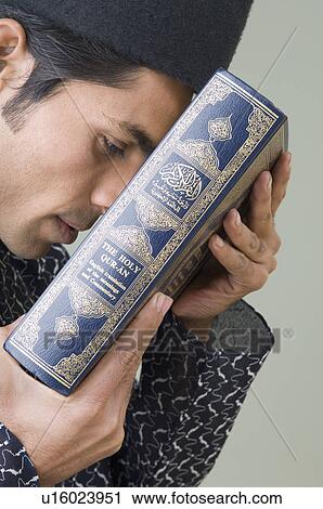 Stock Image - Side profile of a young man holding the Koran against his forehead. Fotosearch