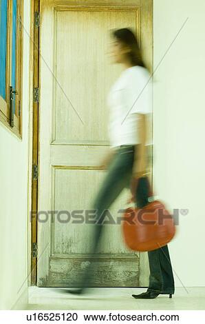 Stock Image - Side profile of a young woman carrying a hand bag and walking out of a room. Fotosearch